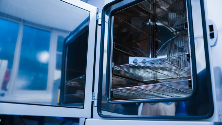 Eine geöffnete, industrielle Temperaturkammer aus Edelstahl mit Blick auf den Innenraum. Auf einem Gitterrost befindet sich ein metallisches Prüfobjekt mit mehreren Verschraubungen, an das Messfühler angeschlossen sind. Im Hintergrund ist die geöffnete Kammertür mit Glasfenster zu sehen.