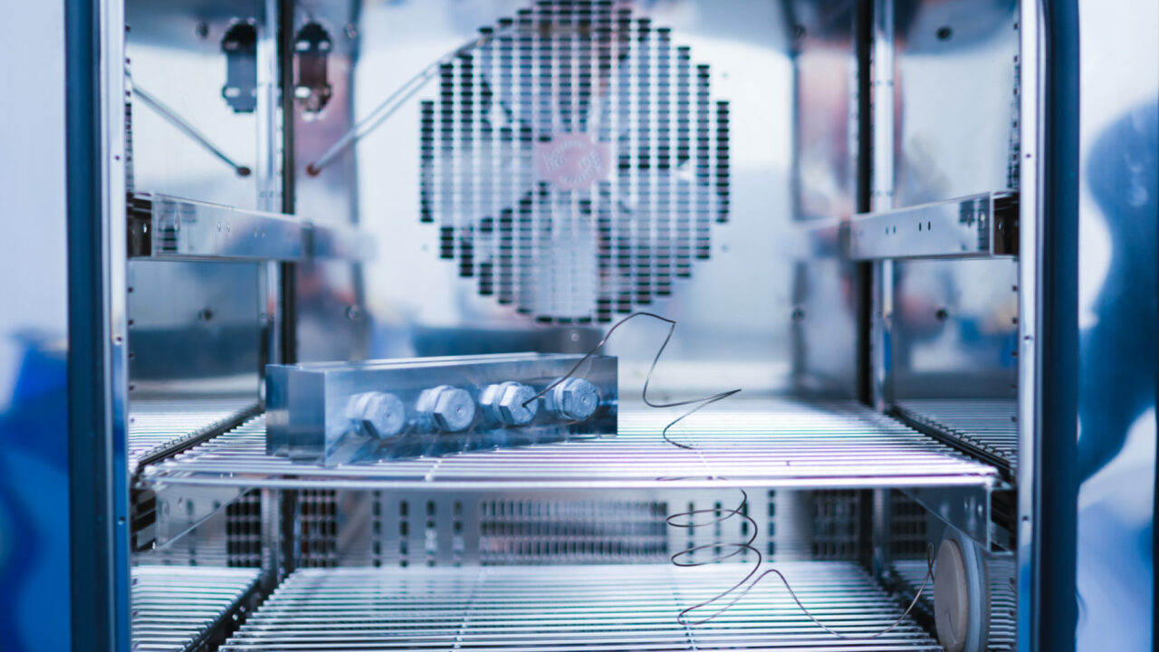 Frontale Nahaufnahme des Innenraums der Temperaturkammer. Ein quaderförmiger Metallblock mit vier großen Sechskantschrauben liegt auf einem Gitterfach. Ein feiner Draht-Sensor führt direkt zu einem der Anschlüsse. Im Hintergrund sorgt ein Ventilator hinter einer Schutzabdeckung für die Luftzirkulation.