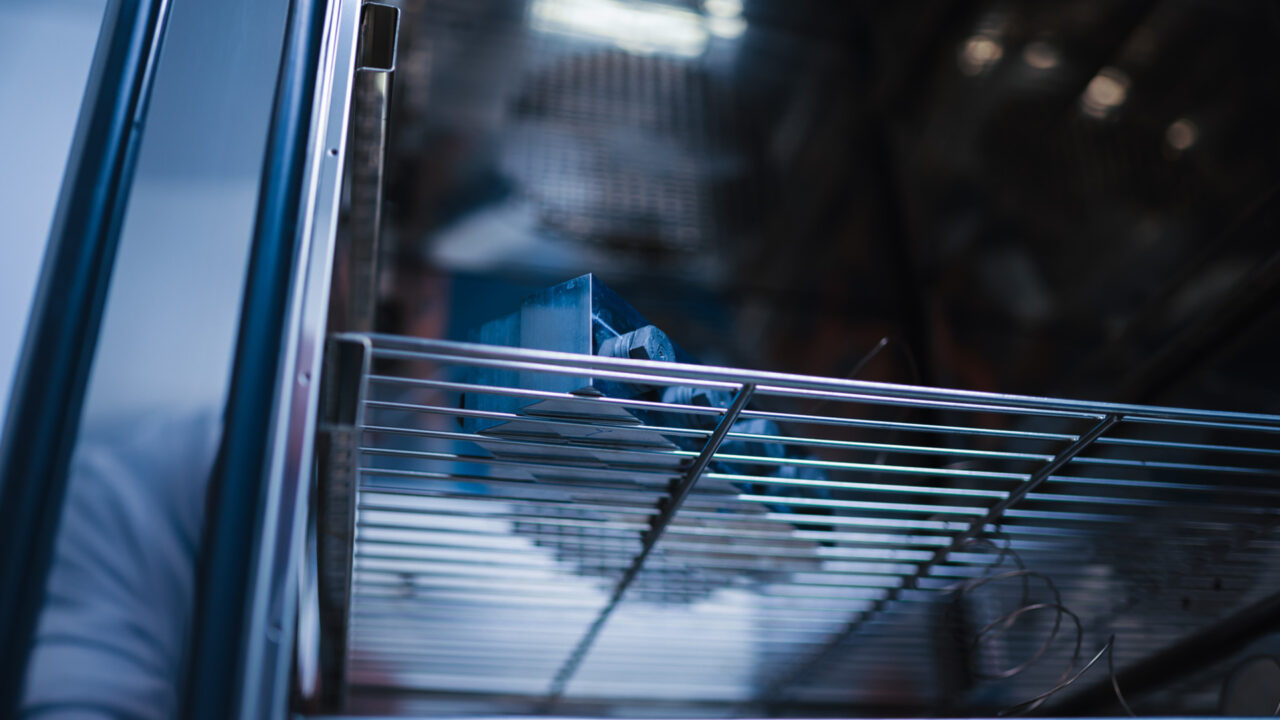 Detailansicht aus einer niedrigen Perspektive auf das Prüfobjekt in der Klimakammer. Der Fokus liegt auf der Gitterstruktur des Fachbodens und den Spiralkabeln der Temperatursensoren, die zum metallischen Testkörper führen.