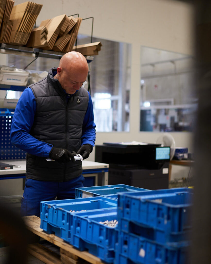 ESKA Mitarbeiter in blauer Arbeitskleidung und grauer Weste steht in einer Lagerhalle. Er trägt Handschuhe und arbeitet mit einem Messwerkzeug (Messschieber) an Kleinteilen. Im Vordergrund stehen blaue Stapelboxen.