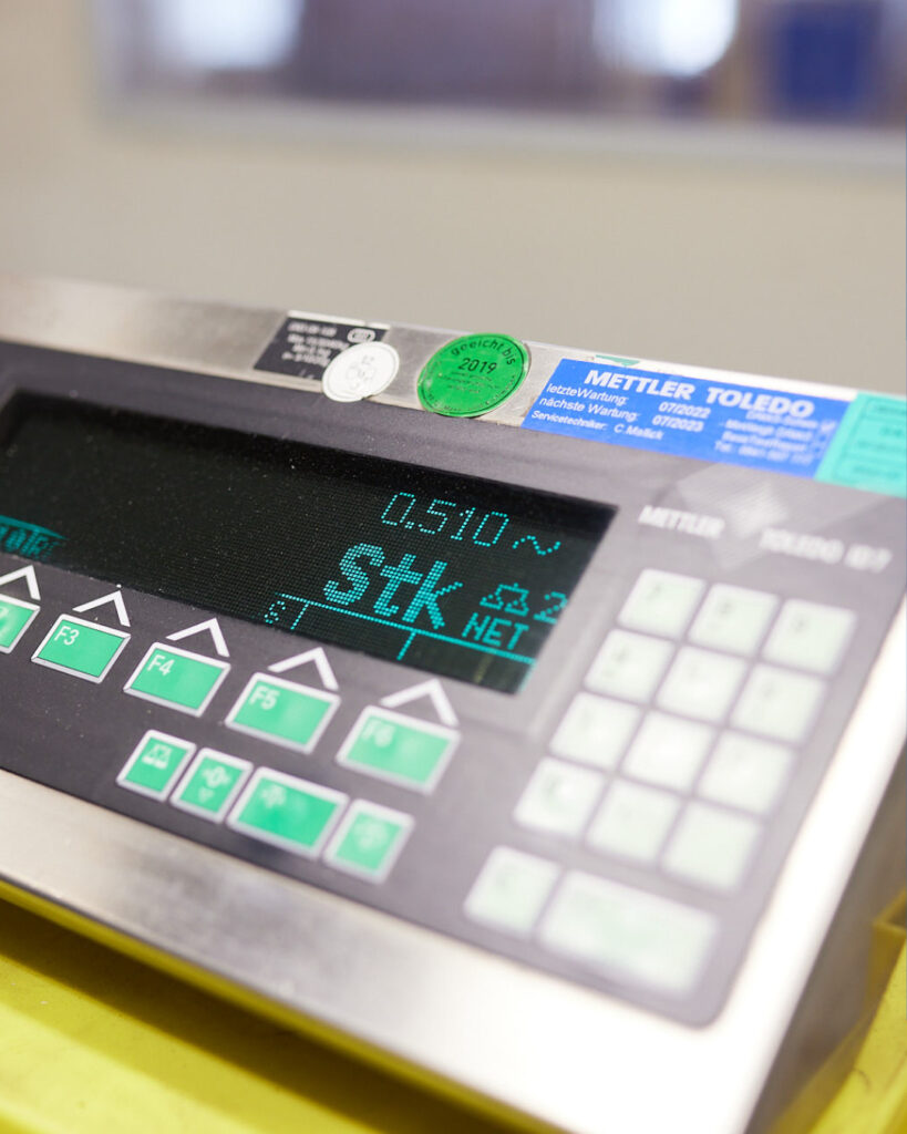 Detailansicht des Displays einer Industriewaage. Die Anzeige zeigt "0.510 Stk NET" in grüner Leuchtschrift. Verschiedene Prüfsiegel sind auf dem Gehäuse sichtbar.