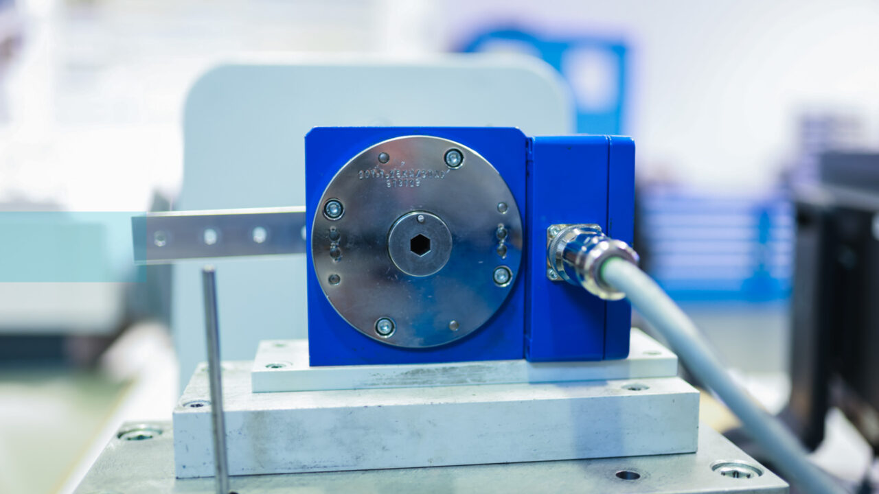 Frontalansicht eines blauen Reibungsprüfstand TesT®. In der Mitte befindet sich eine silberne Drehscheibe mit einer zentralen Sechskantaufnahme. Ein graues Datenkabel ist seitlich mit einer hochwertigen Verschraubung angeschlossen.