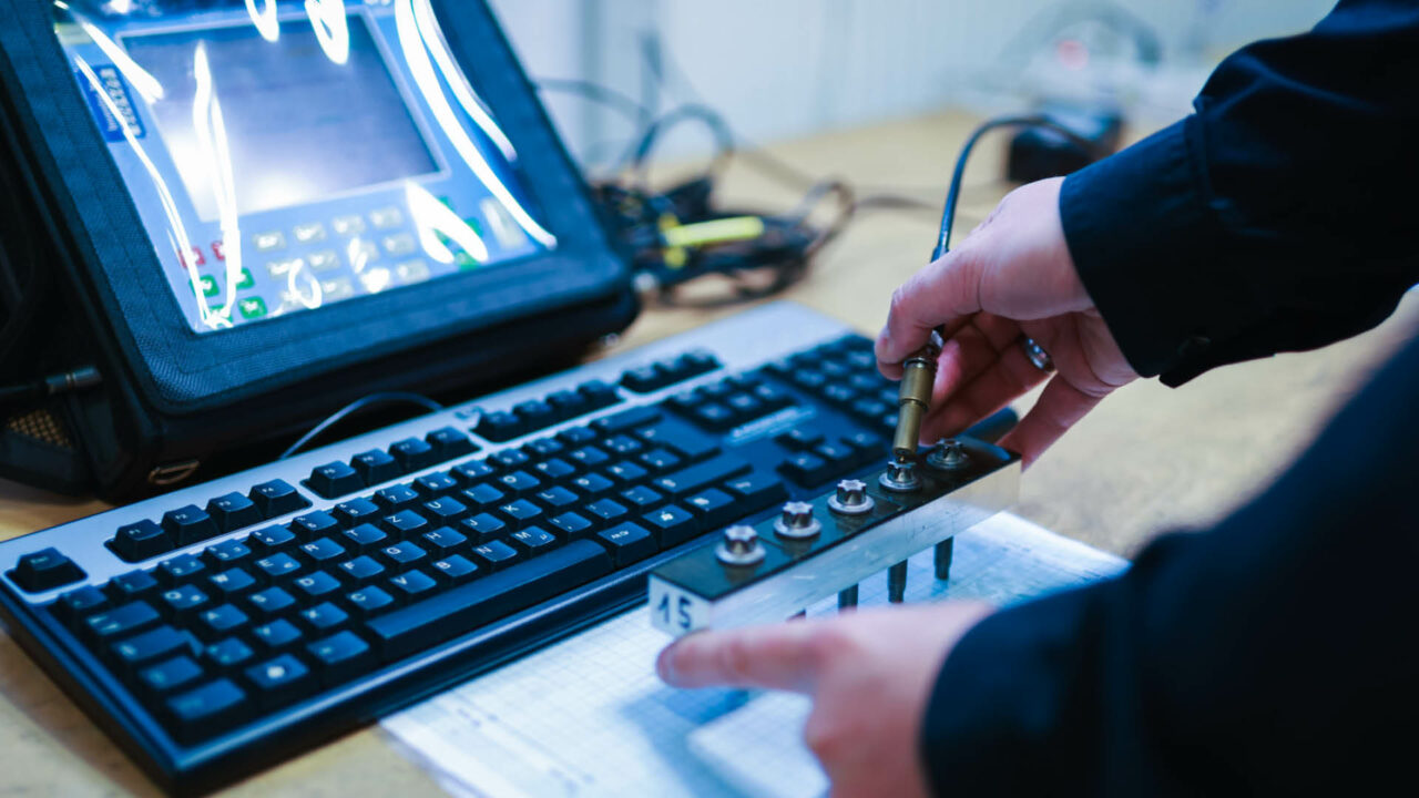 Eine Person führt eine Messung an einer Testvorrichtung mit fünf Torx-Schrauben durch. Mit der rechten Hand wird ein Ultraschall-Messsensor präzise auf den Kopf einer Schraube gesetzt, während die linke Hand die Metallvorrichtung fixiert.