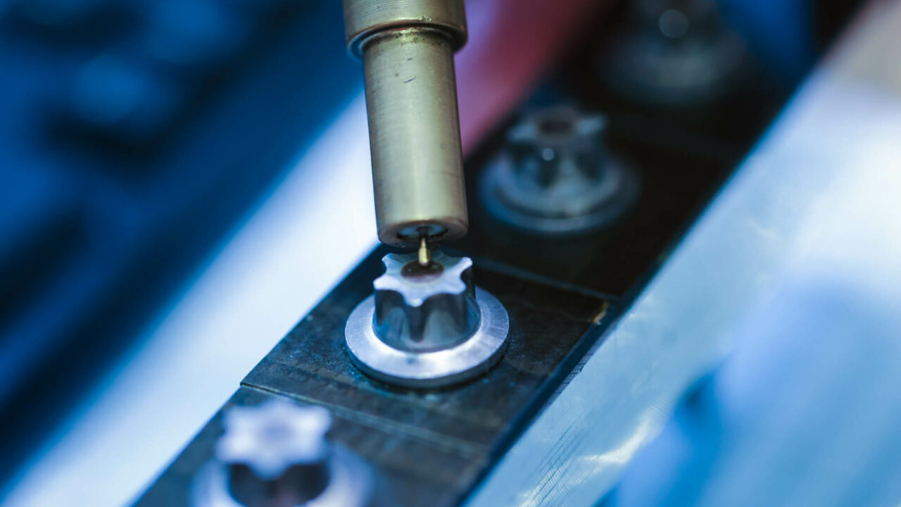 Nahaufnahme eines Ultraschall-Sensorkopfs, der vertikal auf die Mitte eines Torx-Schraubenkopfes aufgesetzt ist. Die Schraube ist Teil einer Testreihe auf einer metallischen Schiene.