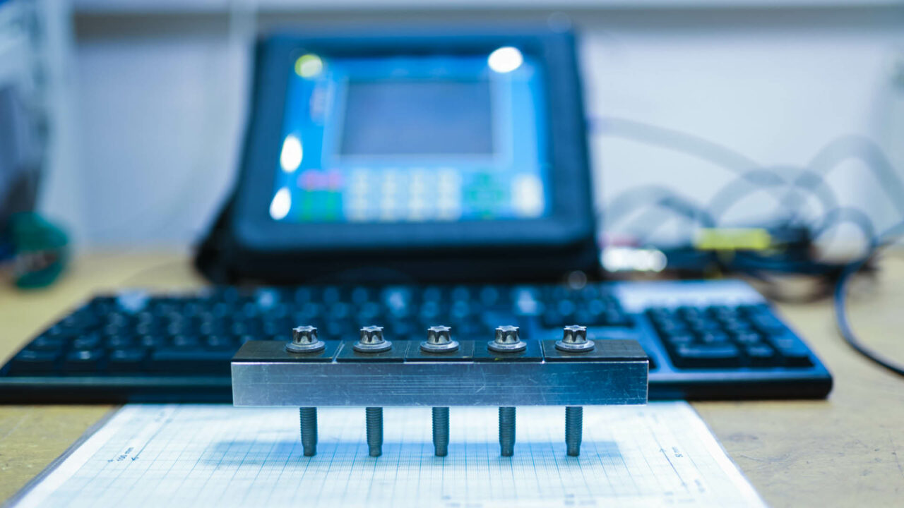 Eine Metallvorrichtung mit fünf montierten Torx-Schrauben steht im Vordergrund auf einem Millimeterpapier. Im Hintergrund stehen eine PC-Tastatur und ein Messcomputer bereit, um die Daten der Vorspannkraftmessung zu erfassen.