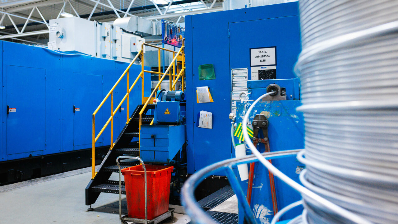 Ein Blick in eine industrielle Produktionshalle mit großen blauen Maschinengehäusen, einer Treppe mit gelbem Geländer und einer großen Kabel- oder Drahtspule im Vordergrund.