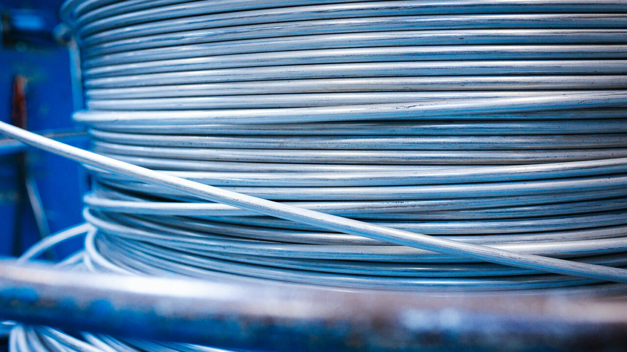Nahaufnahme einer großen, dicht und ordentlich aufgewickelten Spule aus silberglänzendem Metalldraht vor einem blauen Hintergrund.