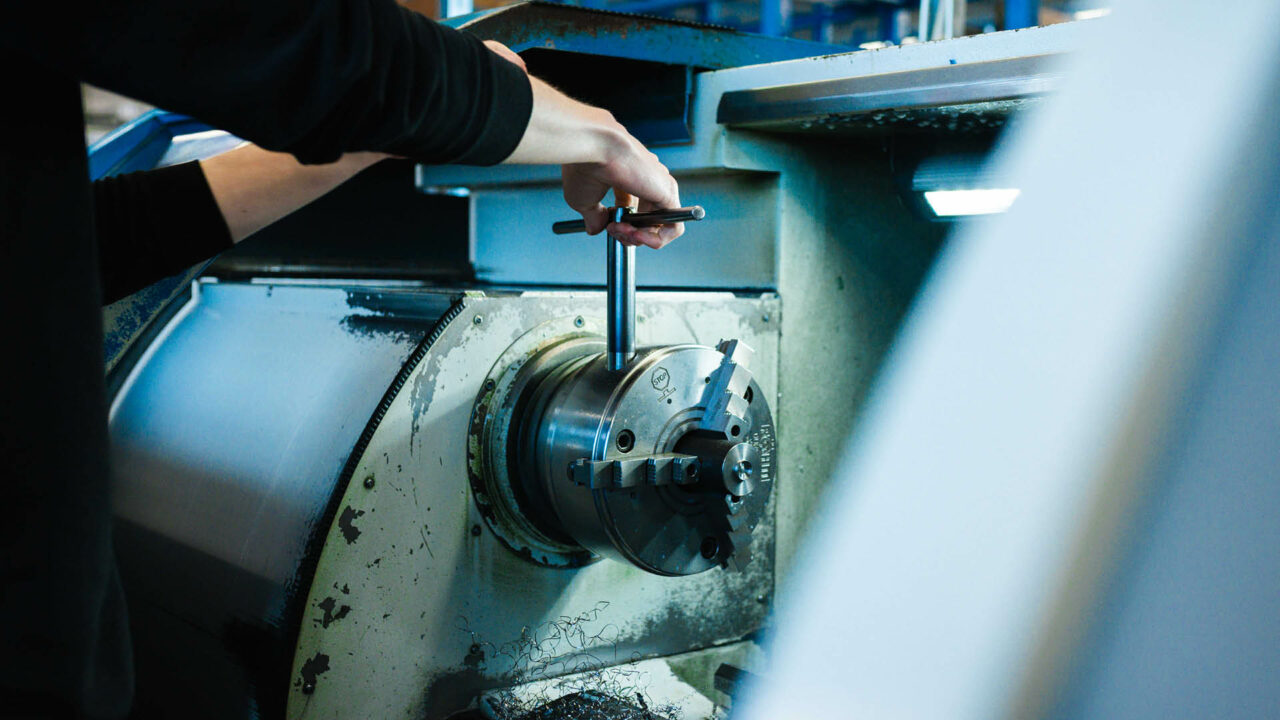 Nahaufnahme der Hand eines Maschinenbediener, der mit einem T-Schlüssel ein Dreibackenfutter an einer Metalldrehmaschine einstellt.