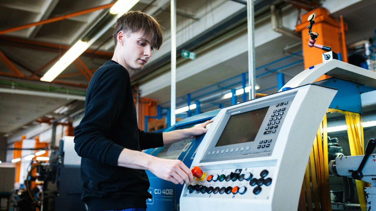 Ein junger Mann im Profil bedient das Heidenhain-Steuerungspanel einer DMT CD 402 CNC-Drehmaschine. Seine linke Hand ruht auf dem Gehäuse, während seine rechte Hand einen Regler auf der Bedienoberfläche einstellt.