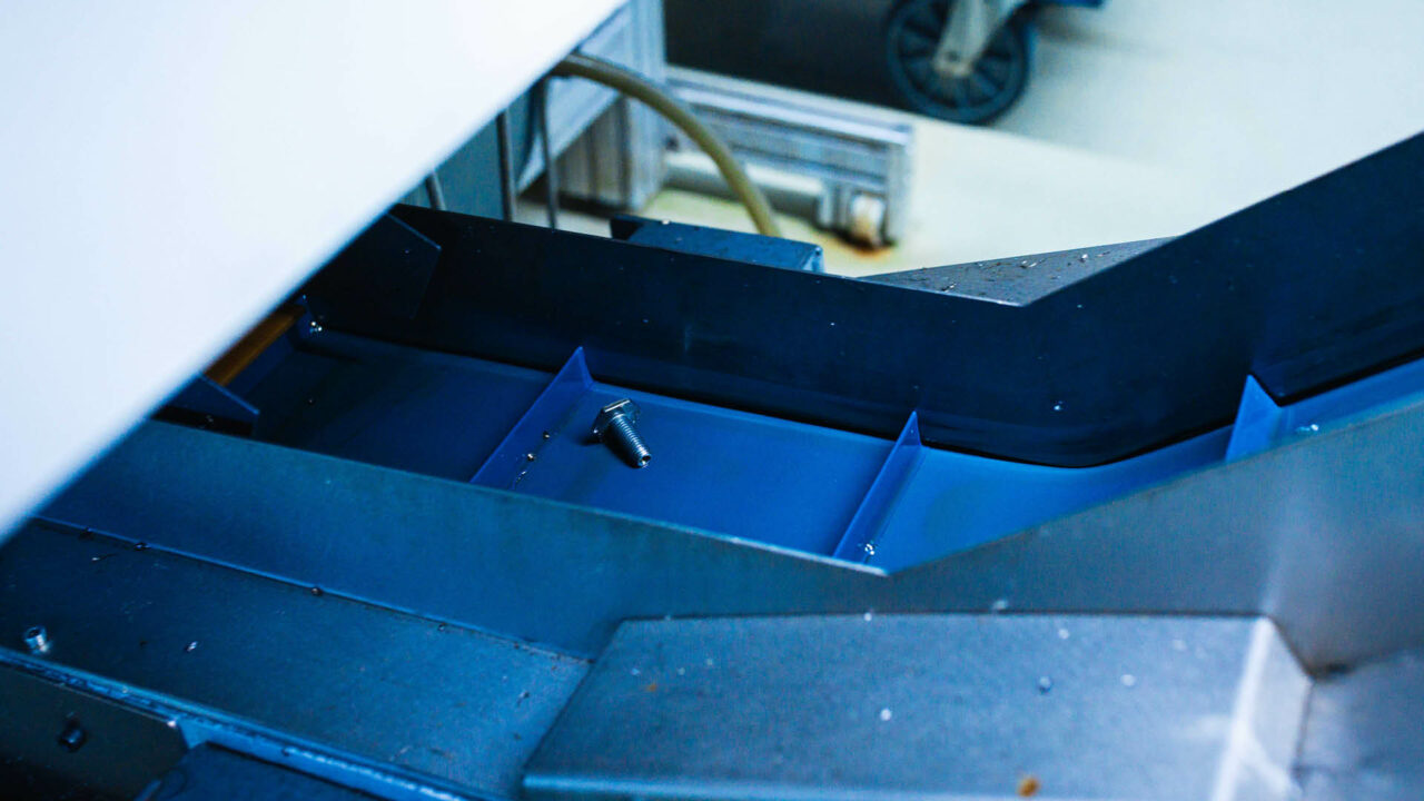 Eine silberne Sechskantschraube liegt mittig in einer blauen, rinnenförmigen Metallschiene einer Industrieanlage.