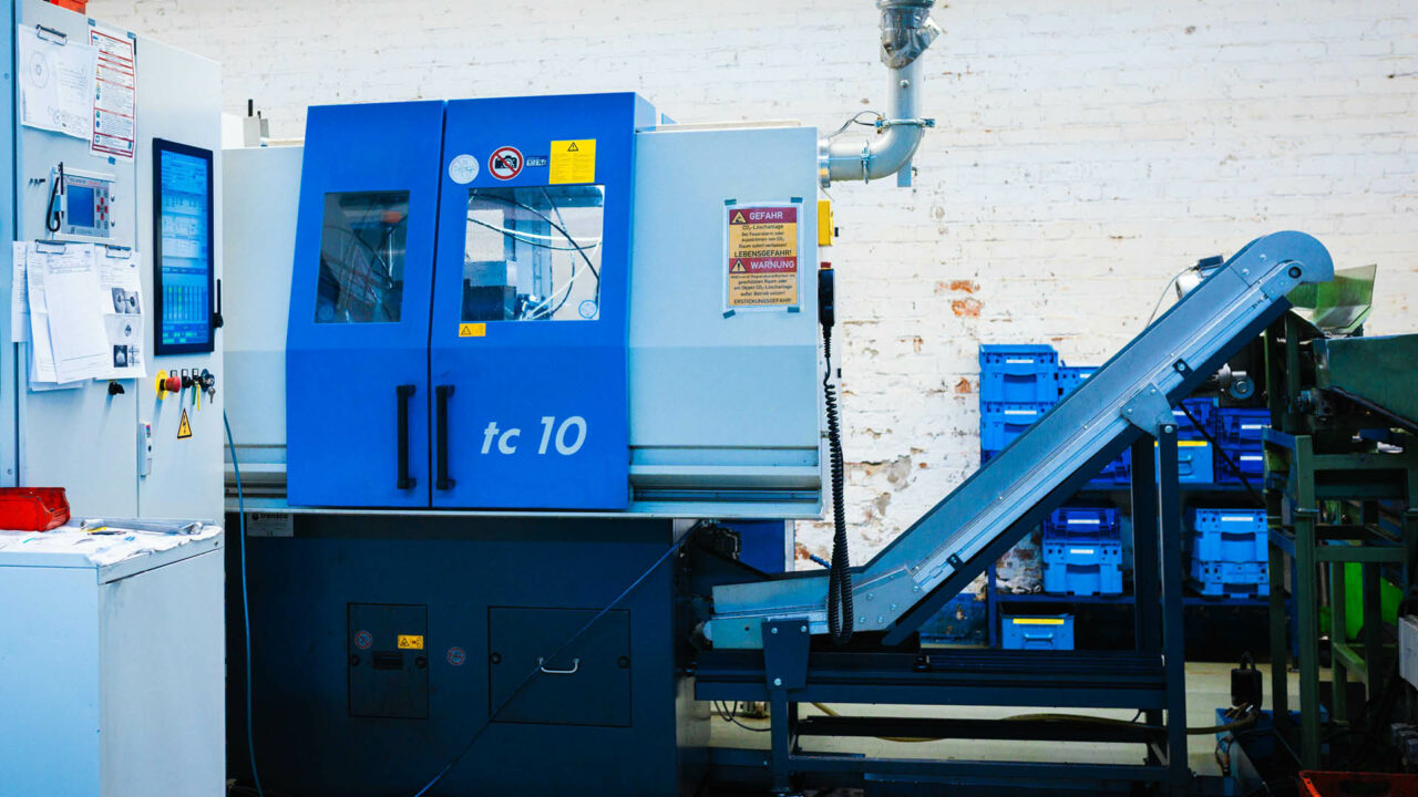Eine blaue und graue Industriemaschine mit der Aufschrift „tc 10“ und Sicherheits-Warnhinweisen. Rechts schließt sich ein ansteigendes Förderband an.