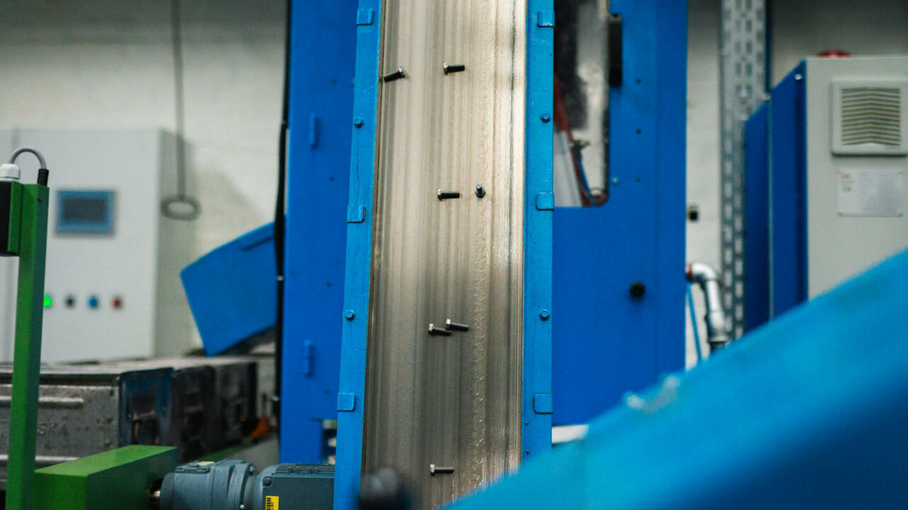 Mehrere kleine, dunkle Schrauben werden auf einem schmalen, senkrecht nach oben verlaufenden Förderband magnetisch mit blauen Seitenwänden transportiert.